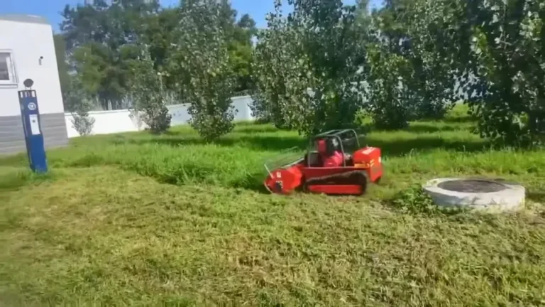 Tracked remote mower cutting grass beside a round drainage access structure